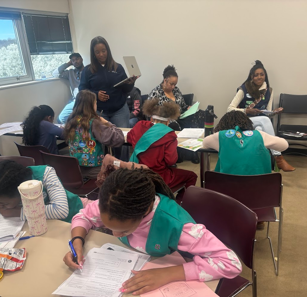 A group of young Girl Scouts seated at tables filling out worksheets during an interactive event.  There is an instructor in the background guiding the activity. 
